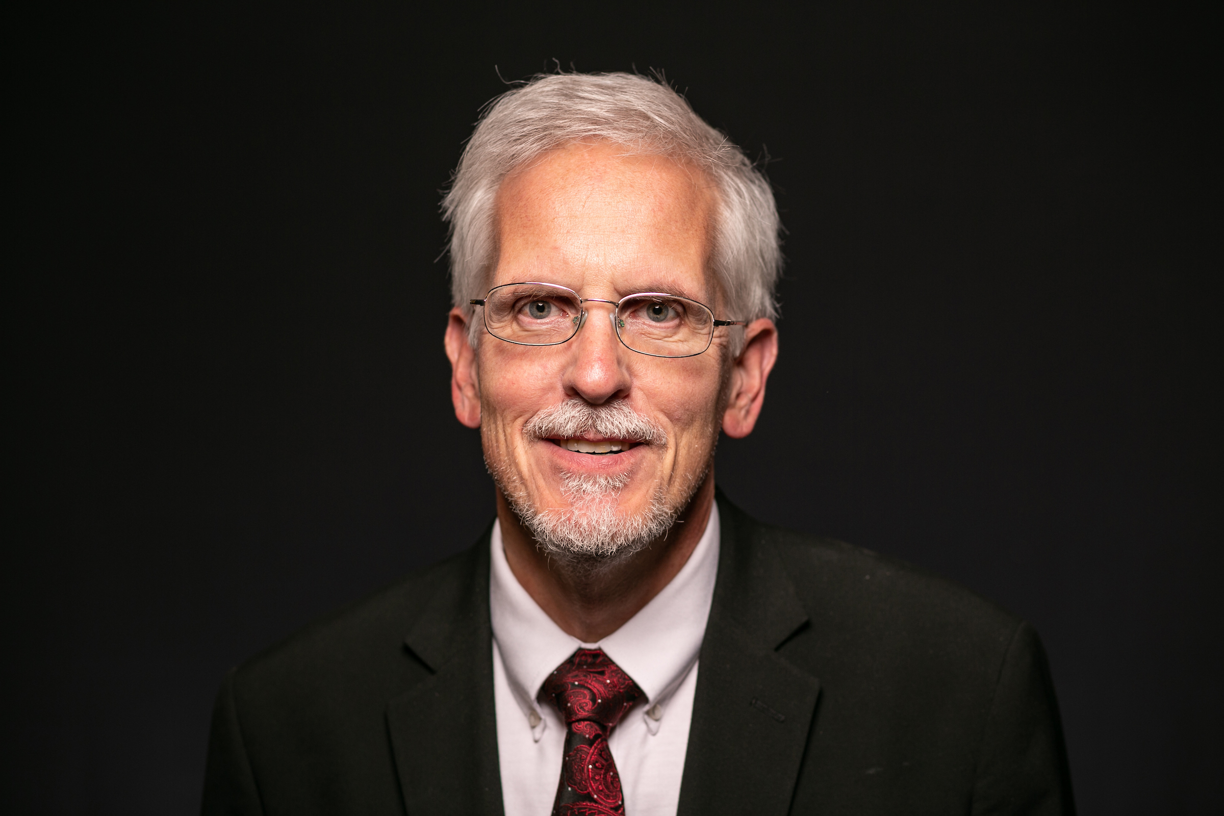 Professional headshot of an older man with glasses and gray hair wearing a suit and tie against a dark background